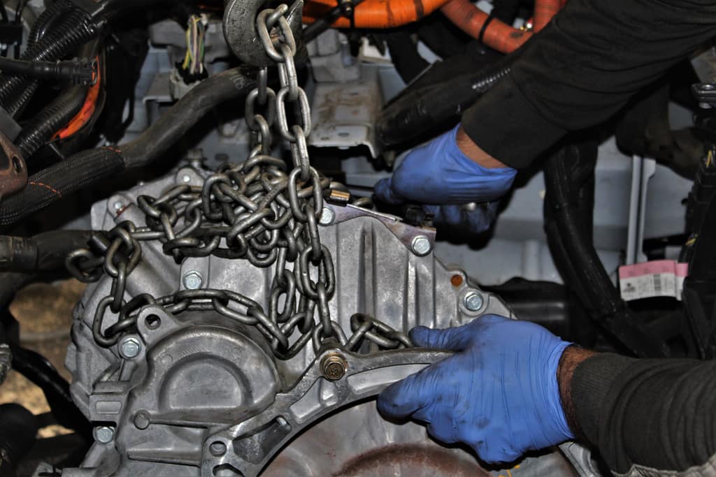 A transmission being chain-hoisted out of a hybrid engine bay, orange high-voltage cables overhead, gloved hands steadying it