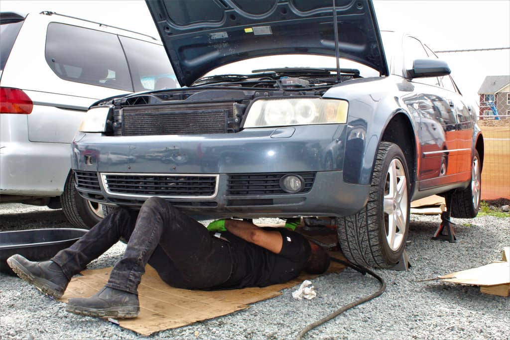 A mechanic lying on cardboard under a dark blue Audi A4 wagon, hood up, in a gravel lot